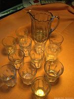 Photo showing one clear Depression glass pitcher and twelve clear glass tumblers arranged on an orange cloth background. Pitcher has Colonial pattern vertical fluted panels and handle.