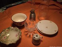Pewter oil lamp with fitted glass chimney, extra glass chimney shown separately, Bavarian China plates, bridal rose mustard jug, crystal compote, Bavarian China bowl, and soup bowl arranged on orange cloth.