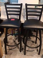 Front view of two black swivel bar stools showing seats, backs, and legs with footrests.