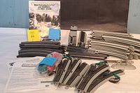 Wide photo showing extensive collection of grey and black curved and straight model railroad tracks, track switches, accessories, and the instruction manual in the background.