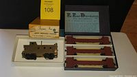 Great Northern brass caboose in white tray with original box, alongside box containing three Northern Pacific log flats in packaging with label.