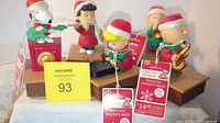 Five Hallmark Peanuts characters (Snoopy, Schroeder, Lucy, Charlie Brown, Linus) in Christmas attire with instruments on rectangular wood-tone bases, all bearing tags