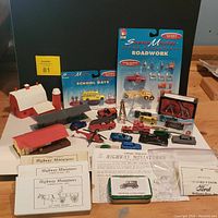 Photo showing unopened blister packs labeled SceneMaster roadwork and school days with various miniature vehicles and figures in front, multiple miniature buildings including barns and station, and several sealed unassembled miniature car kits with instructions.