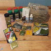 Photo showing entire set of landscape scenery supplies on wooden table including rubber rocks base, multiple packets and bottles, clump foliage, sealer, and flock containers.