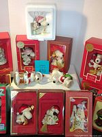 Photo showing 10 boxed Lenox Snoopy china ornaments, some out of boxes and some in red boxes, various holiday themed designs.