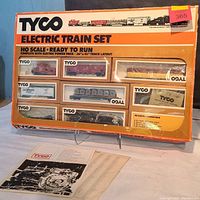 Photo of Tyco Electric Train Set #7308 in original box with orange and white packaging. Clear display windows show multiple train cars including freight boxcars, tanker car, coal car, and locomotive. The box is in vintage condition with some corner wear. Instruction sheets placed in front.