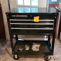 Front view of the black U.S. General rolling tool box with two drawers and bottom shelf, containing spools of soldering wire and a bag of supplies on the lower shelf.