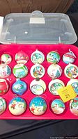 Top view of open plastic storage case holding 20 vintage Snoopy Christmas ball ornaments, in compartments, various festive colors and designs