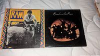 Two vinyl LP albums placed side by side on a white fabric surface: Paul McCartney Ram with bright colorful cover and Wings Band On The Run with dark cover showing band members in poses.