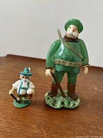 Photo showing the pair of figurines, hunter and mini beer drinker, side by side on wooden surface.