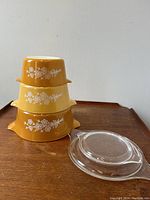 Three vintage Pyrex casserole dishes stacked including two lids beside them, featuring the Butterfly Gold white floral pattern on gold backgrounds.