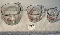 Three Anchor Hocking Fire-King measuring cups (1, 2, and 4 cups) displayed on a white textured cloth with red measurement markings visible.