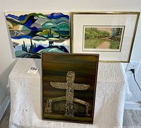 Three wall artworks displayed on a white textured surface. Left ceramic tile artwork with colorful mountain and sailboat scene, center front wood-mounted etched metal with Thunderbird and LongHouse totems, right framed Michaela Davidson print of a river scene.