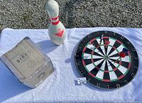 Photo showing the set consisting of the bowling pin, Bingo game box, and dartboard with darts laid on white cloth.