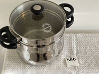 Stainless steel multi cooker pot with steamer insert and glass lid sitting on a white textured cloth, showing front and side views of the cooker with black handles.