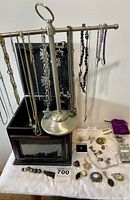 Photo showing large black jewelry box with clear glass front and a silver-tone metal necklace stand holding various necklaces. Assorted jewelry including brooches, rings, earrings, and a watch are displayed on table.