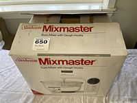 Original retail box of Sunbeam Mixmaster Euro-Mixer with Dough Hooks, predominantly white box with product image and branding, showing the box mostly closed with some tape still intact.