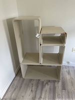 Four wooden cube shelving units stacked in various configurations, showing front and side views. The wooden cubes are painted off-white with scratches and dings. The floor is wood laminate and the wall is plain white.