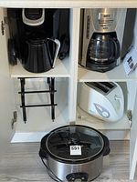 Kitchen cabinet with Keurig coffee maker and black thermos, Black & Decker drip coffee maker, Betty Crocker toaster, and Crock Pot slow cooker inside.