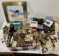 Large metal tin filled with assorted costume jewelry including pendants, bracelets, earrings, pins, and necklaces with decorative motifs and colorful accents.