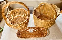 Three wicker baskets on a white table: large round basket with thick handle, oval basket with handle, and long narrow basket in front.