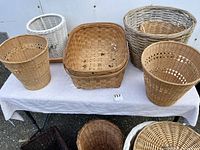 Ten wicker and woven baskets arranged on and under a white cloth-covered folding table outdoors against a white wall and truck trailer background.