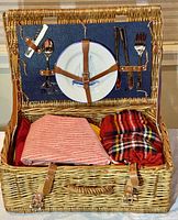 Open wicker picnic basket showing interior contents including two ceramic plates, metal utensils, and two folded cloth items.