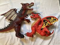 Two large hard plastic dragon toys, one red with yellow and black wing patterns, one dark red with beige wings, posed on a white cloth.