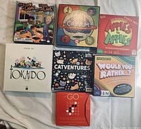 Seven board games arranged on a white surface showing front box art: The Floor is Lava, Cranium, Apples to Apples, Tokaido, Catventures, Would You Rather, and The Go Game.