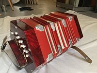 Angled side view showing red concertina body, white buttons, bellows and leather strap