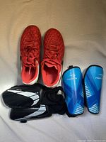 Top view of the red kids' soccer cleats paired with a set of black and white shin pads with elastic straps visible, showing the size 1 cleats and one set of shin pads.