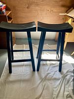 Photo showing two matching black wooden stools with curved seats and four legs with support bars, placed on a white cloth on wooden floor.