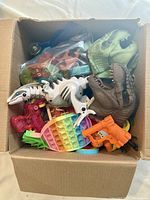 Top view of a cardboard box filled with various toys including dinosaur figures, hand puppets, a rainbow fidget popper, a toy dart gun, and other small items.