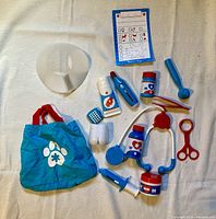 Photo of various plastic veterinary play medical tools, including a cone collar, stethoscope, syringe, thermometer, scissors, medicine bottles, and a patient chart, with a blue bag with paw print.
