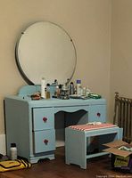 Bright blue vanity with red knobs and attached round mirror. Matching seat with red and white cushioning. Items on vanity surface include bottles and tubes of personal care products. Some scuffs on top.