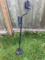 Full view of black metal detector standing on grass, showing long shaft, coiled wire, armrest and round search coil.