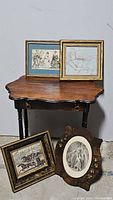 Four antique framed prints arranged on and around a dark wood table; various frame styles including ornate gold and carved wood, images include period clothing and horse racing.