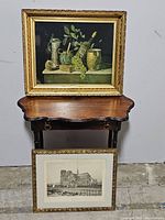 Overall view of both framed art pieces, still life painting on a table and Notre Dame print resting below.