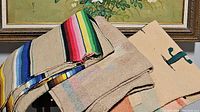 Image showing three folded Southwestern style blankets on a wooden surface under framed artwork. One blanket has multicolored horizontal stripes with fringes visible.
