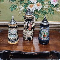 Three King German beer steins displayed side by side on a wooden table, showing different designs and pewter lids.