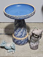 Concrete bird bath with blue basin and decorative leaf pattern shown with green frog statue and owl decoy.