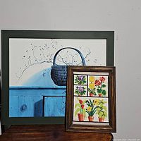 Photo showing both framed artworks with blue dresser painting in background and smaller floral needlepoint in front.