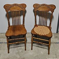 Pair of two carved oak spindle back dining chairs side by side. One chair has a woven seat cover. Chairs show carved decorative crest rails and turned spindles on back .