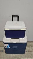 Blue Rubbermaid cooler on top of large blue Coleman wheeled cooler with white lids.