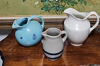 Three pitchers together showing different shapes and colors on wooden surface under a painting.