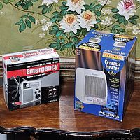 Both boxed items placed on wooden furniture with floral wallpaper background: Eton FR300 Emergency Radio and Pelonis HC-441W Ceramic Heater.