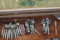 Photo of mixed assorted flatware including knives, forks and spoons on a wooden surface beneath a framed floral painting