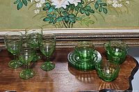 Full display of the lot showing 4 green glass stemmed wine glasses, 3 green glass bowls, and 4 cups with saucers in green glass.