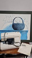 Kenmore off-white sewing machine with attached cords and foot pedal on wooden surface, manual beside it, blue basket painting in background.