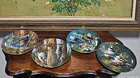 Four decorative bird plates displayed on a wooden table, showing detailed bird artwork and gold rims.